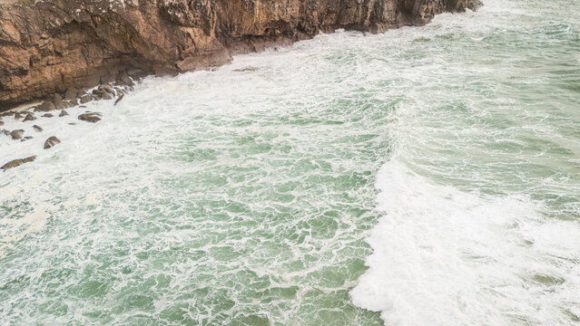 Aerial view of rough turquoise sea waves crashing against the steep rocky cliffs of Sagres with white foam and spray Sagres, Faro, Portugal.