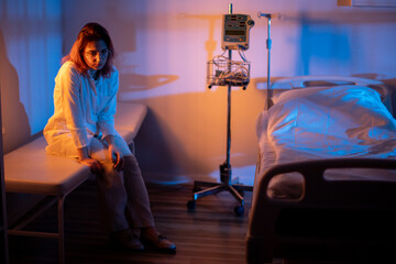 Solitary person resting on bed within dimly illuminated ward setting. Lone patient sits on bed as faint blue glow and orange lighting create contrasting atmosphere © fotodrobik
