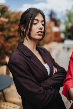 Young woman in a dark blazer stands with crossed arms and a dismissive expression. Outdoor portrait captures a confident professional in a casual urban setting.