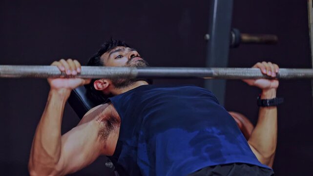 Muscular fit man lifting heavy barbell during intense bench press exercise in modern dark gym for chest strength training workout showing extreme focus building power