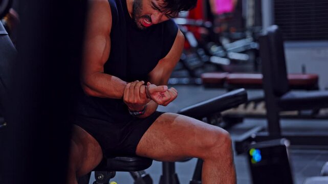 Fit man sitting on bench inside dark gym feeling intense pain holding wrist injury experiencing deep physical struggle after heavy muscle workout and exhausting daily fitness exercise routine