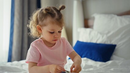 Fototapeta premium Curious toddler in pigtails wearing a pink shirt, sitting on a bed and playing with a toy in a bright, cozy bedroom, exploring and learning during early childhood development