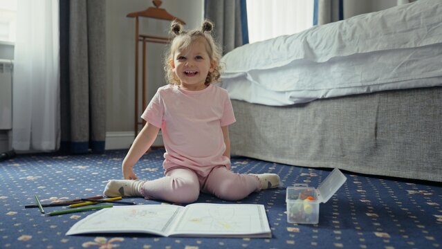 Little girl sitting on bedroom carpet coloring in a book with colorful pencils, smiling and focused in a cozy indoor scene of playful creativity and childhood joy