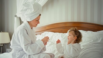 Fototapeta premium Mother and daughter wearing bathrobes and towel turbans, applying moisturizer on their faces together during a relaxed morning skincare routine on a white bed