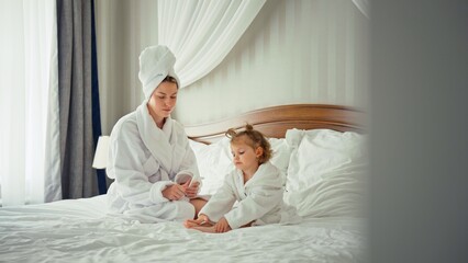 Fototapeta premium Mother and daughter wearing white bathrobes, with mom's hair wrapped in a towel, sitting on a comfortable bed in a light filled bedroom, sharing a calm moment