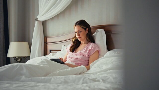 Young woman in pink pajamas relaxing in a luxurious bed, reading an e reader tablet with white sheets, enjoying a quiet morning of cozy leisure and digital entertainment at home or hotel