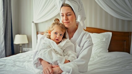 Obraz premium Mother hugs young daughter, both wearing white bathrobes and feeling comfortable in a hotel room, symbolizing family bonding, care, and wellness during a vacation