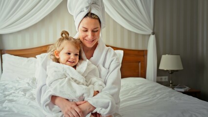 Fototapeta premium Mother and daughter wearing cozy white bathrobes and towels, smiling and embracing while sitting on a comfortable bed in a bright bedroom, starting a relaxed morning routine together