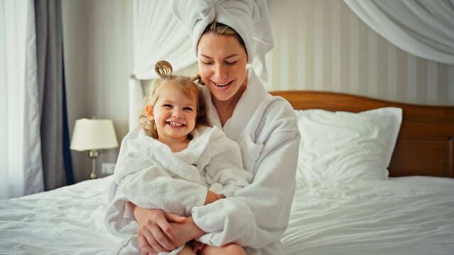 Mother and daughter in bathrobes sitting on bed after shower, smiling and hugging in a cozy bedroom moment of family love, care and morning togetherness