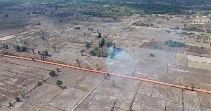 Aerial view of dry, segmented farmland crossed by a dirt road, with a small patch of smoke rising from the center.