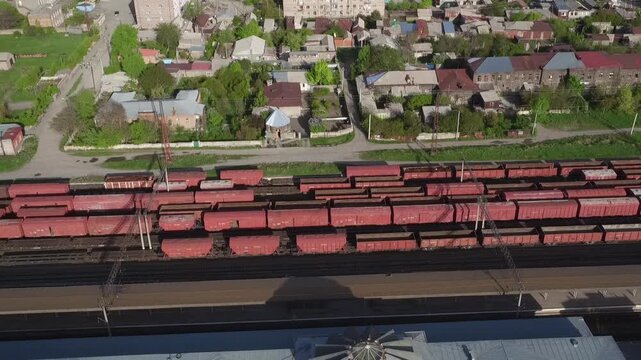 High angle aerial drone view of a railway station with red cargo wagons parked on tracks, surrounded by residential buildings in a small town during sunny spring day.

