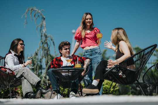 A group of teenage friends sitting and standing together outdoors enjoying drinks and conversation. Young people relax on a sunny patio in casual summer outfits sharing a social moment.