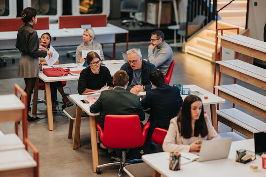 A diverse business team collaborates around a table in a modern coworking space. Colleagues discuss documents and laptops during a focused group meeting.