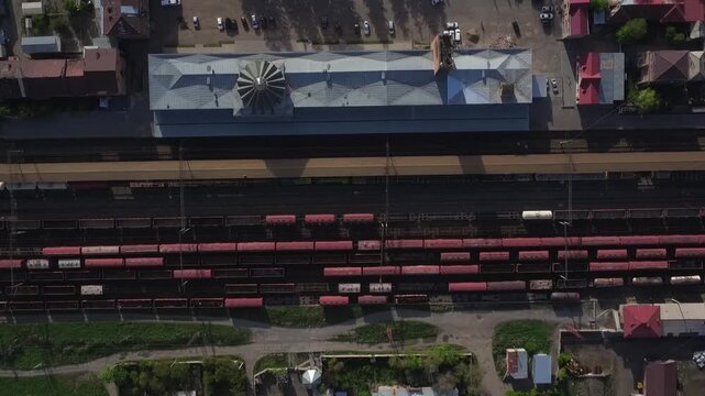 High angle aerial drone view of a railway station with red cargo wagons parked on tracks, surrounded by residential buildings in a small town during sunny spring day.

