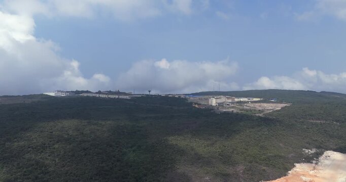 Backward aerial view of a sprawling hilltop resort complex, including the Bokor Hill Station in the background, nestled in a vast, forested landscape under a cloudy sky.
