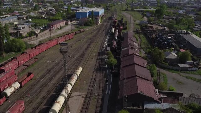 High angle aerial drone view of a railway station with red cargo wagons parked on tracks, surrounded by residential buildings in a small town during sunny spring day.

