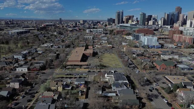 City core progression frames Denver beyond broad community block near West 11th Ave where residential streets shift toward larger civic buildings. Sunlit roofs green pockets, park playground, CO