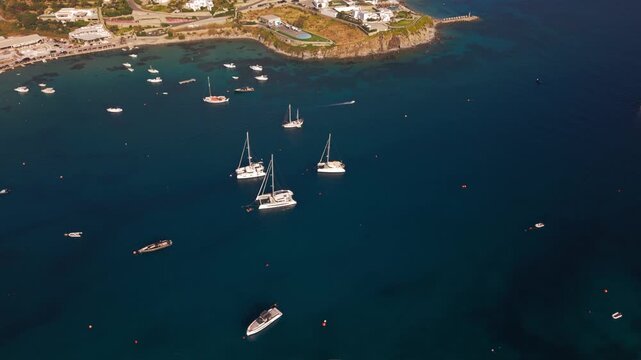 High angle panoramic drone footage of Ornos beach and bay in Mykonos island, Greece. Luxury yachts and boats anchored in the turquoise Aegean sea surrounded by traditional whitewashed villas.