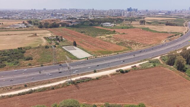 Aerial drone view of busy highway cutting through agricultural fields with dense urban skyline in Hod Hasharon, Israel

