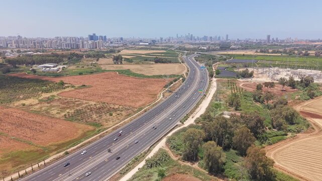 Aerial drone view of a major highway stretching toward Tel Aviv skyline through agricultural fields near Hod Hasharon
