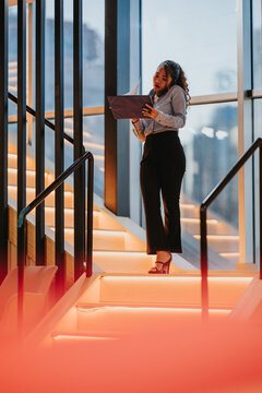 Businesswoman standing on lit stairs reviewing a folder and speaking on a mobile phone. The professional reviews paperwork on illuminated office steps near large windows.