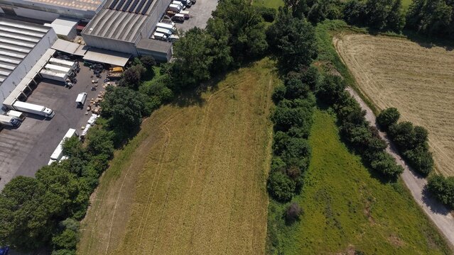 Industrial Edge Meets Nature: An aerial view captures the stark contrast between a sprawling industrial complex with loading docks and parked vehicles, and an adjacent patch of untamed greenery.