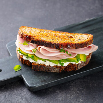 Healthy sandwich with ham, avocado and soft white cheese on toasted whole grain bread on a black marble board. Grey stone background. Close up.