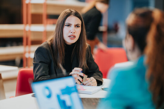 A businesswoman speaks with a concerned expression while discussing documents across a table. Colleagues engage in a tense business meeting in a contemporary office setting.