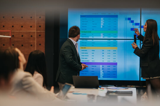 Business professionals present a project schedule on a large screen to colleagues in a meeting room. A presenter points to timelines and charts while teammates review the plan together.