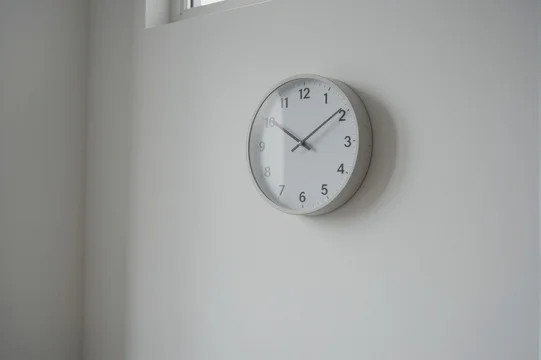 A minimalist analog wall clock with a white dial and light gray frame is mounted on a plain white wall in a modern residential setting.