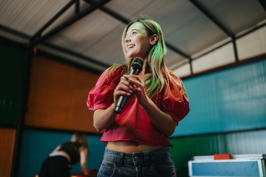 A young woman holds a microphone and smiles while performing with friends. Casual indoor karaoke or rehearsal scene showing friends enjoying music together.