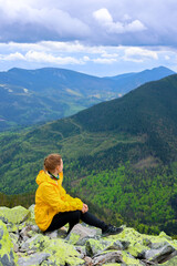 Fototapeta premium Woman hiking in mountains. Woman in yellow on the rocky mountain peak on the background of green mountain forests. Green tourism, hiking concept. Close up woman hiking. Hiking tourism advertisment. 