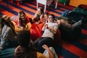 Fototapeta premium A group of young friends laugh and relax on bean bags in a colorful lounge. They are friends enjoying casual conversation and drinks in a cozy, playful setting.