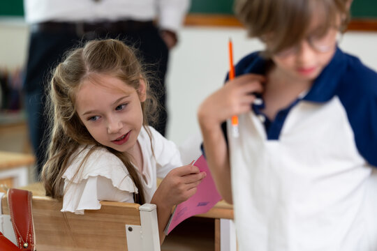 Elementary girl smiles while classmate holds pencil and worksheet near desk, back to school classroom activity builds friendship, participation, study confidence, peer learning, social skill.