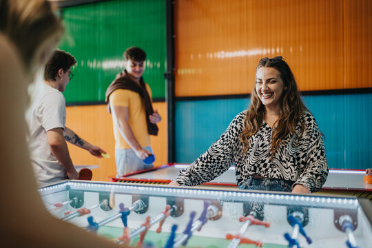 A group of friends play foosball and arcade games, smiling and enjoying themselves. Casual young adults socialize around a foosball table in a bright, colorful game room.