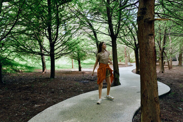 Fototapeta premium Woman walking on winding path through forest park among tall trees and nature, casual outfit with jacket tied at waist, looking up during a relaxed outdoor stroll.