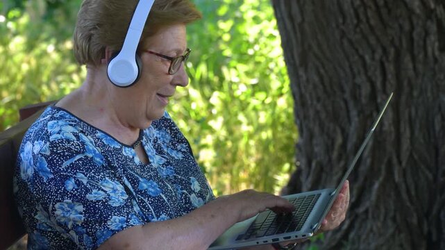 Senior woman in a video call wearing headset sitting in a bench in the park.