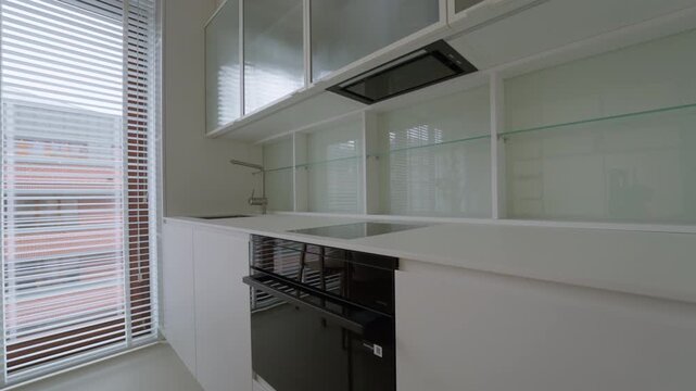 Built in black oven and induction cooktop under extractor hood inside brand new renovated vacant apartment room with empty white kitchen cabinet counter after interior remodeling finish.