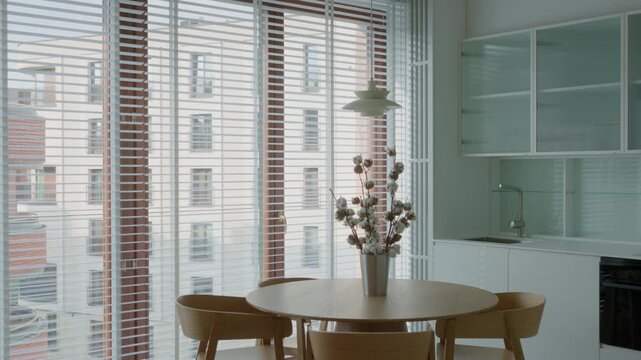 Dining room table with chairs and cotton flower vase stands inside empty kitchen interior. Vacant apartment residential property contains window blinds house accommodation real estate asset.