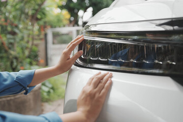 Close-up of hands touching the headlight and hood of a white car outdoors, checking vehicle condition or cleaning with soft daylight and blurred garden background. © Yanisa