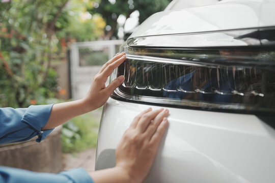 Close-up of hands touching the headlight and hood of a white car outdoors, checking vehicle condition or cleaning with soft daylight and blurred garden background.