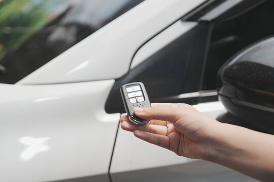 Close-up of a hand holding a car key fob near a white vehicle door, suggesting remote unlocking or entry, modern transportation technology concept.