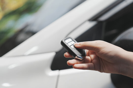 Close-up of hand holding car key fob near white vehicle door, pressing remote button for locking or unlocking, shallow depth of field, modern transport and security concept.