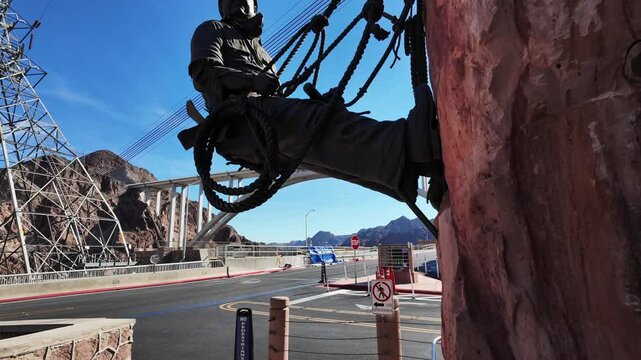 The Hoover Dam on the Colorado River looking at a statue of a construction worker hanging from the rock with the Dam Bridge in the Background
