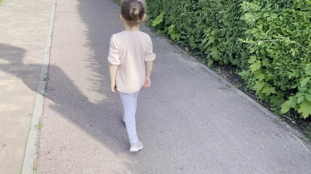 Little girl walking alone along a path near green bushes, back view