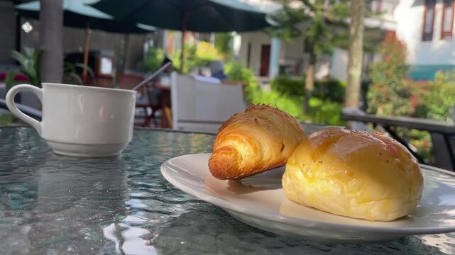A close-up shot of a white cup and two pastries on a glass table in an outdoor cafe setting, with umbrellas and greenery in the background.