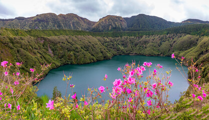 Green volcanic crater lake Lagoa de Santiago in Sete Cidades, Sao Miguel Island, Azores, Portugal. Lush mountains and pink flowers surround the scenic emerald lagoon water. © Max Maximov