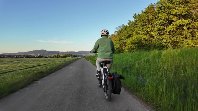 Woman cyclist riding electric bicycle on rural road in evening countryside  