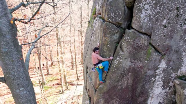 Top view of man climbing natural stone wall above forest floor, using rope protection. Scenic aerial footage for fitness, determination, challenge, and adventure travel themes