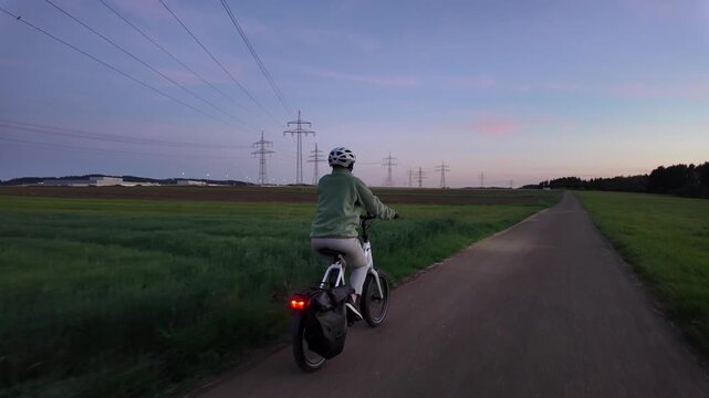 Woman cyclist riding electric bicycle on rural road in evening countryside  